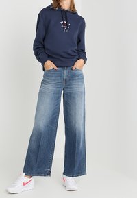 Navy hoodie with a logo on the chest, paired with wide-leg light blue jeans featuring a light wash and frayed hem. White sneakers with pink accents.