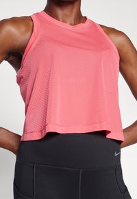 Personne athlétique portant un crop top en maille corail sans manches et un legging noir taille haute Nike, les mains sur les hanches.