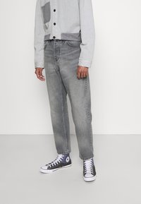 Jasnoszare jeansy z denimu o prostym kroju nogawek, z wyblakłymi miejscami. Do tego szaro-czarne trampki za kostkę z widocznymi białymi sznurowadłami.