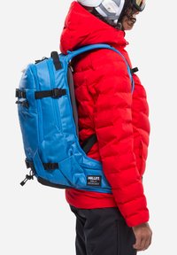 Sac à dos bleu avec des sangles latérales et des boucles, porté sur une veste matelassée rouge. Comprend une étiquette de marque sur la partie inférieure du sac. Texture lisse.