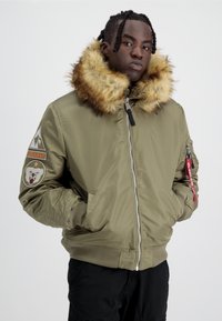 Giubbotto bomber verde oliva con collare in pelliccia sintetica, chiusura con zip e polsini a costine. Presenta dettagli con toppe sulle maniche e una tasca laterale.