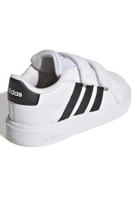 Bílé kožené tenisky s černými třemi pruhy, suchými zipy, texturovanou bílou gumovou podrážkou a hladkým černým akcentem na patě.