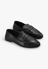 Mocasines de cuero negro con un acabado suave, que presentan un diseño plisado en la puntera y un sutil detalle de correa en la parte superior. Suela plana.