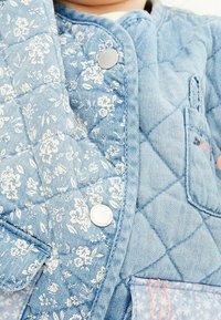 Jeansjacke mit einem hellblauen Blumenmuster, gestepptem Material, Druckknopfverschluss und einer Tasche mit kontrastierenden Nähdetails.