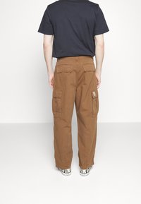 Eine Person trägt braune Cargo-Hosen mit mehreren Taschen, ein schwarzes T-Shirt und weiße Converse-Sneaker mit Zebra-Streifen und steht mit dem Rücken zur Kamera.