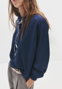 Maglietta a maniche lunghe blu navy con una vestibilità morbida, caratterizzata da un collo rotondo e una sciarpa blu a fantasia annodata al collo. Abbinata a pantaloni grigi.
