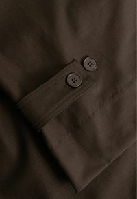 Smeđi materijal glatke teksture, sa manžetom na dugmad koja ima dva tamna dugmeta, vidljivi detalji ojačanih šavova na ivicama.