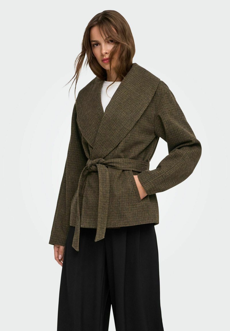 Femme portant une veste portefeuille marron à carreaux avec un large col et une ceinture, associée à un pantalon noir à jambes larges, debout devant un fond uni.