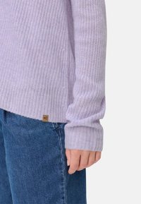 Lila gestrickter Pullover mit gerippter Textur und langen Ärmeln, der ein kleines Markenetikett nahe dem Saum aufweist, kombiniert mit blauen Denim-Jeans.