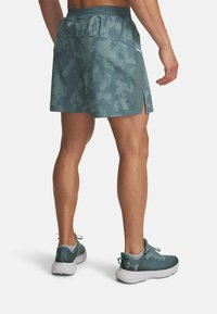 Bermudas deportivas para hombre en color azul verdoso, con un patrón de camuflaje, aberturas laterales y cinturilla elástica, combinadas con zapatillas de correr a juego en azul verdoso.