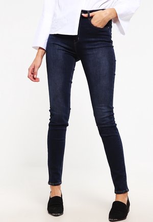 Vrouw met donkerblauwe skinny jeans, wit shirt met lange mouwen en zwarte instapschoenen, staand met één hand in de zak.