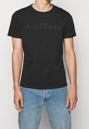 Mann trägt ein schwarzes John Richmond T-Shirt mit verblasstem Logo und hellblauen Jeans, steht vor einem einfarbigen Hintergrund.