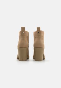 Botines de ante beige con tacones de bloque, tiradores acanalados y cuellos de tela texturizada, fotografiados desde la parte trasera sobre un fondo blanco.