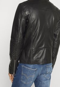 Černá kožená bunda s prošívanými kruhovými detaily na rukávech, zipovými manžetami a rovnými liniemi. Kombinováno s modrými džíny.