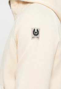 Ανοιχτό μπεζ φούτερ με υφασμάτινη υφή, με ραμμένο μαύρο σήμα λογότυπου στον αριστερό ώμο και κουκούλα με φερμουάρ.