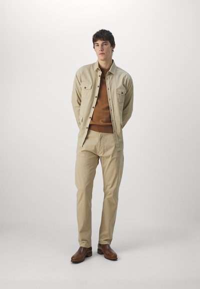 Beige shirt jacket with two chest pockets, worn over a brown crewneck sweater and beige pants; brown leather shoes complete the outfit.