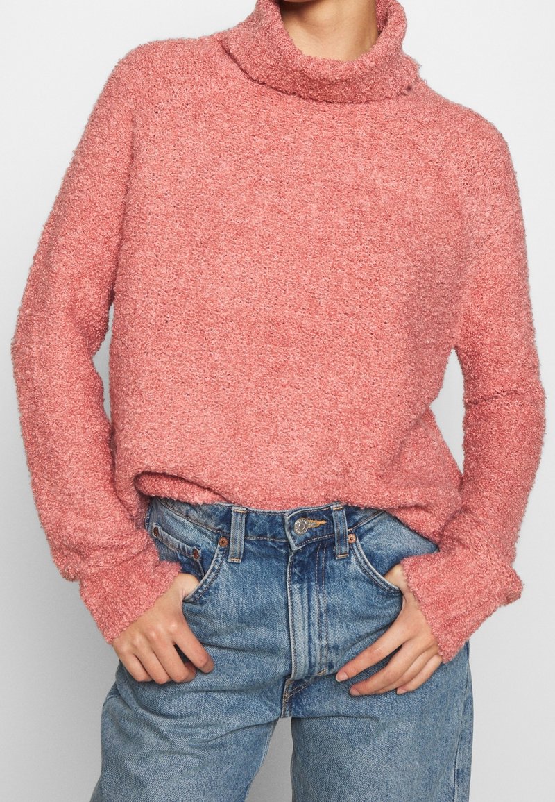 Pull en maille texturée rose avec col montant et manches longues, associé à un jean en denim bleu taille haute.