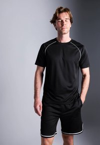 Jovem em camisa atlética preta e calções, com os olhos fechados e uma mão no bolso, contra um fundo cinza bicolor.