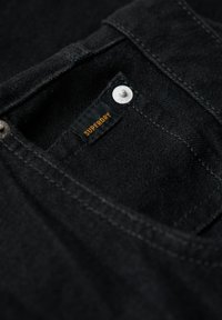 Tissu en denim noir avec une finition texturée, comportant une petite étiquette indiquant "SUPERDRY" en orange, et un détail de bouton-pression argenté.