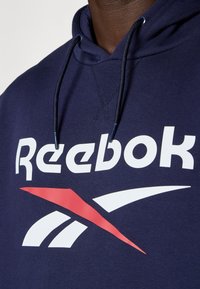 Hanorac albastru marin cu un mare logo alb Reebok și accente roșii. Material din bumbac, cu glugă cu șnur și detalii cusute.