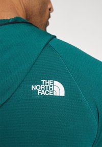 Teal hoodie with a textured fabric, featuring a white logo on the back, designed with a hood and raglan sleeves for enhanced fit.