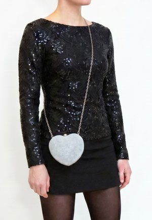 Femme portant un haut noir à manches longues pailleté et une jupe, tenant un sac en forme de cœur argenté avec une bandoulière chaîne traversant son corps.