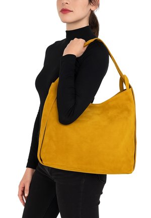 Borsa a tracolla gialla in suede con una texture morbida, ampia tracolla e design minimalista, tenuta sotto il braccio contro una maglia nera.