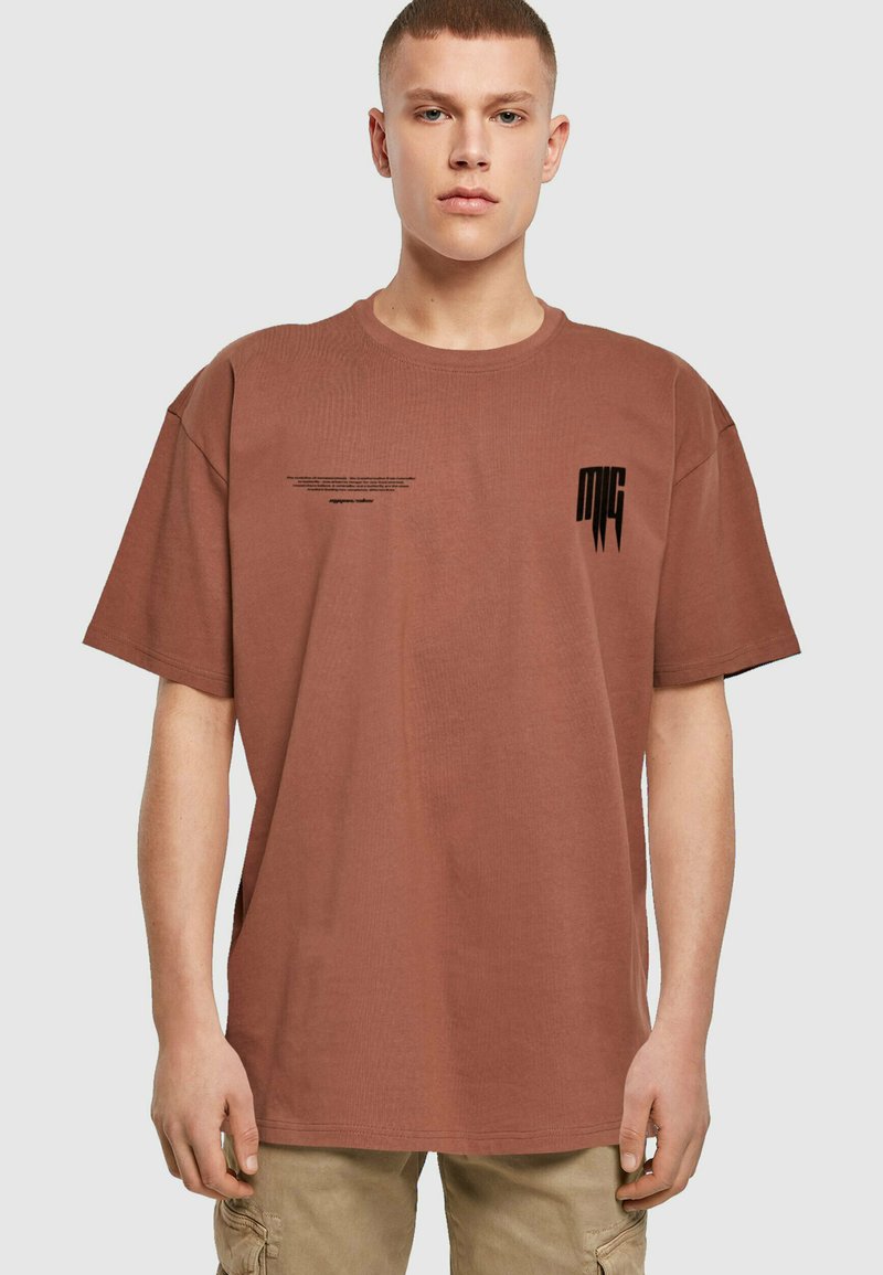 Brun bomulds t-shirt med korte ærmer. Har et lille sort grafik på venstre bryst og tekstdetaljer over den øverste del af kroppen.