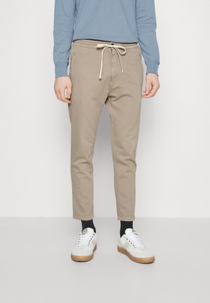 Osoba ubrana w beżowe spodnie ze ściągaczem, czarne skarpetki, białe trampki oraz niebieską koszulę z długim rękawem, stojąca na jednolitym tle.