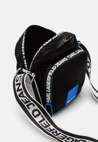 Črn najlon crossbody nahrbtnik z glavnim predelkom na zadrgo, modrim poudarkom in nastavljivim trakom z ponavljajočim se vzorcem belega logotipa.