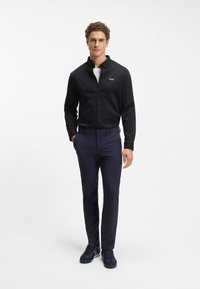 Μαύρο πουκάμισο με κουμπιά και διακριτικό λογότυπο, συνδυασμένο με σκούρο μπλε παντελόνι στενής εφαρμογής και σκούρα αθλητικά παπούτσια, με μοντέρνα γραμμή και καθαρές γραμμές.