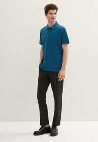 Türkiz színű póló ing gallérral és rövid ujjakkal, sötétszürke slim-fit nadrággal és fekete fűzős cipővel párosítva.