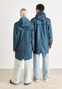Jachete impermeabile de culoare teal, cu glugă, textură netedă și ventilație în spate, asociate cu blugi de tip wide-leg de culoare albastru deschis. Disponibile în două mărimi.