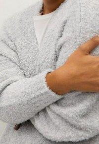 Grauer, flauschiger Cardigan aus strukturiertem Stoff. Mit einem Schalkragen und Knopfdetails. Die Ärmelabschlüsse sind umgeschlagen und zeigen das weiche, gemütliche Material.