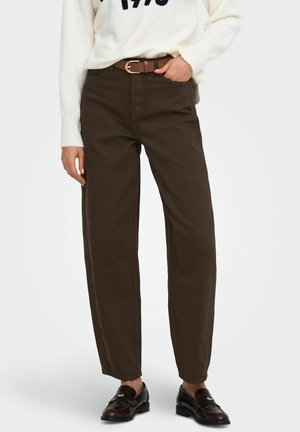 Personne portant un pantalon taille haute marron foncé avec une ceinture marron, un pull blanc et des mocassins marron foncé, debout avec une main dans la poche.