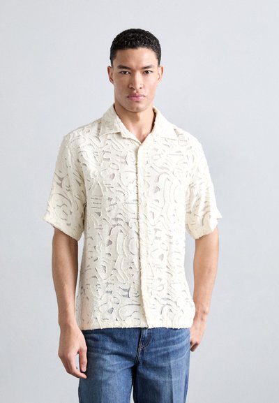 Chemise à manches courtes à boutons couleur crème, ornée de motifs abstraits texturés, avec un col et une coupe décontractée, associée à un jean bleu.