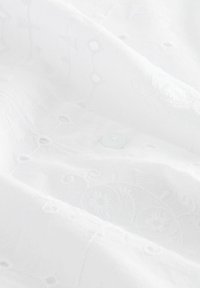Weißer Stoff mit komplizierten, tonal abgestimmten Stickmustern und kleinen Ausschnitten. Ein einzelner transparenter Knopf ist unter den Falten sichtbar.