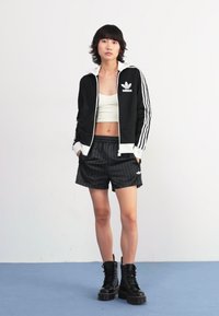 Svart adidas-träningsjacka med vita ränder, vit crop top, svarta rutiga shorts och svarta plattformstövlar. Enfärgad bakgrund.