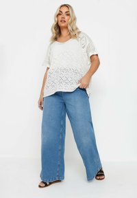 Top de ganchillo blanco con mangas cortas, combinado con unos jeans de pierna ancha azules. El conjunto presenta un corte relajado y un patrón de calado.