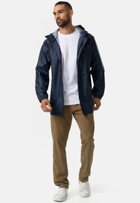 Marine regenjas met capuchon en grijze voering, verstelbare trekkingstrengen, ritssluiting aan de voorkant en elastische boorden, gecombineerd met beige broek en witte sneakers.