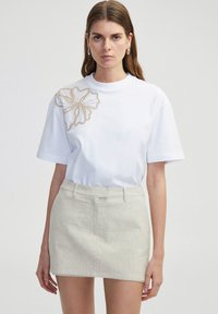 T-shirt en coton blanc avec une grande fleur beige brod�ée sur l'épaule gauche, associée à une jupe beige clair. Manches courtes, coupe décontractée.