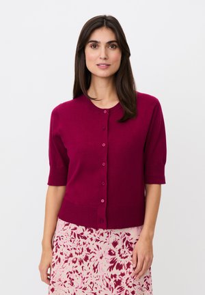 Donna con capelli scuri e lisci che indossa un cardigan magenta a maniche corte abbottonato e una gonna rosa con motivi floreali scuri, in piedi su sfondo bianco.