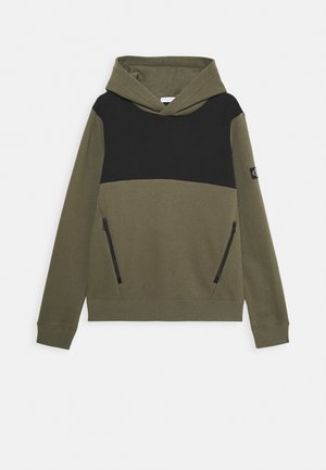 Sudadera de color verde oliva con un panel superior negro, presenta bolsillos laterales con cremallera y un dobladillo acanalado, hecha de un tejido suave y texturizado.