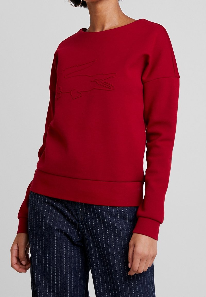 Red sweatshirt made of soft fabric, featuring a textured crocodile logo on the front, round neckline, and ribbed cuffs and hem.