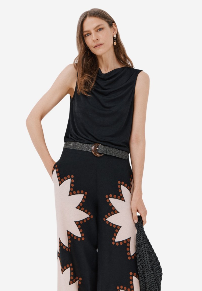 Top nero senza maniche con scollatura drappeggiata, abbinato a pantaloni larghi con sfondo crema e grandi motivi a esplosione marrone.