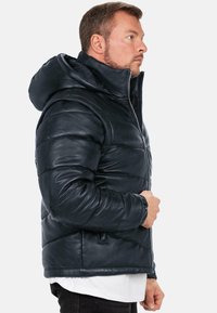 Giacca di pelle nera imbottita con zip frontale e un ampio cappuccio. Presenta polsini a coste e tasche laterali per una maggiore funzionalità.