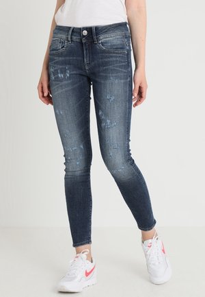 Jeans ajustados en denim oscuro con detalles desgastados, diseño de cinco bolsillos, cintura de altura media, y combinados con zapatillas blancas con acentos en rosa.