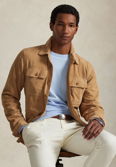 Giacca marrone chiaro con bottoni e due tasche sul petto, indossata sopra un maglione a girocollo azzurro cielo, abbinata a pantaloni color crema.