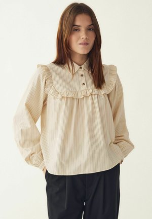 Bluzka w kremowe paski z falbanianym kołnierzem, długimi rękawami i zapięciem na guziki. Materiał ma gładką fakturę z delikatnym połyskiem.