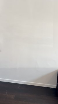 Bijela zidna površina s blagom teksturom, minimalnim oznakama, uz tamni drveni pod. Utikač je vidljiv s lijeve strane.